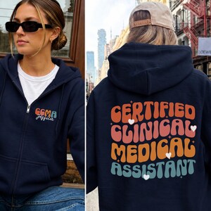 May include: A navy blue zip-up hoodie featuring a white t-shirt underneath. The front left chest displays the text CCMA Jessica. The back features the text CERTIFIED CLINICAL MEDICAL ASSISTANT in a retro, wavy font with small white heart accents.