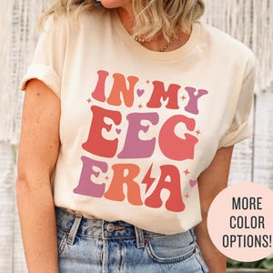 Puede incluir: Una camiseta de color crema con el texto "In My Egg Era" en una fuente retro con colores rosa, naranja y morado. El texto está rodeado de pequeños corazones y un rayo.