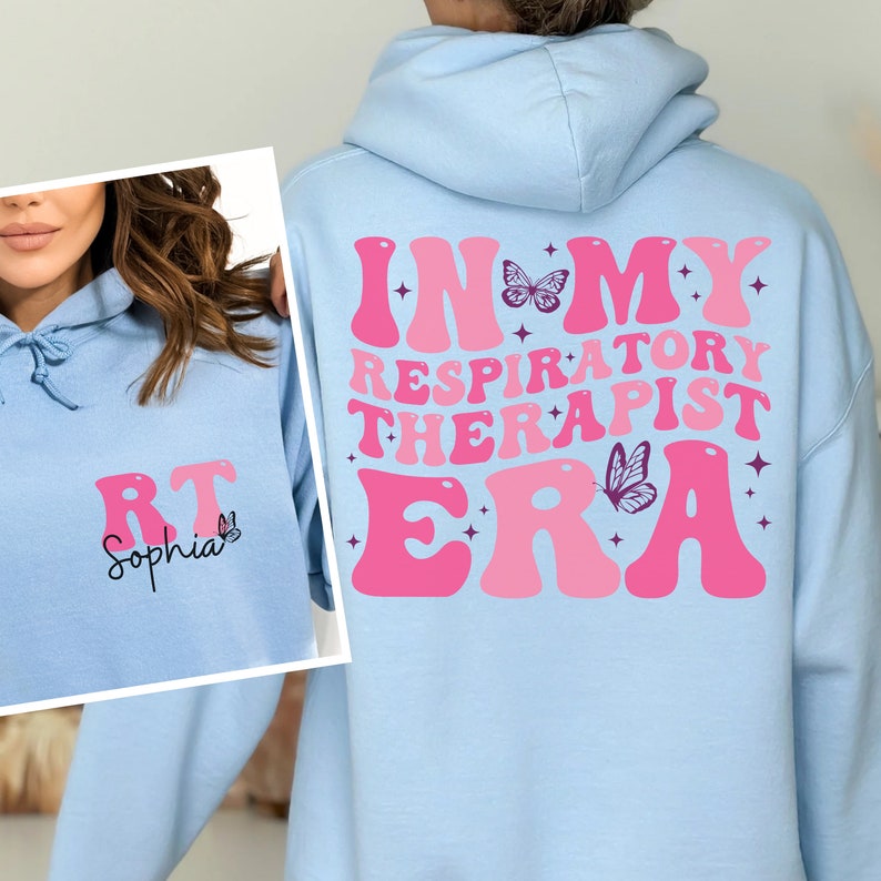 May include: Light blue hooded sweatshirt with the text "In My Respiratory Therapist Era" in pink retro-style lettering with butterflies and stars. The sweatshirt also has the text "RT Sophia" on the front.