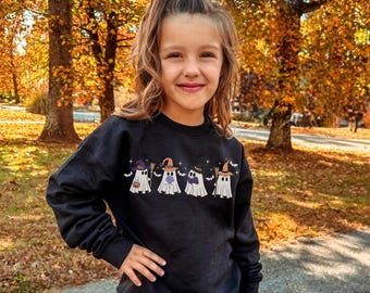 Puede incluir: Una sudadera negra con una fila de fantasmas de dibujos animados que llevan sombreros de bruja y sostienen una escoba, una pajarita y una linterna. Los fantasmas son blancos con contornos negros. El fondo muestra a una persona joven frente a árboles con follaje otoñal.
