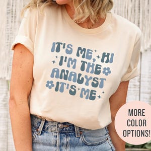 May include: A white t-shirt with the text "It's Me, Hi I'm The Analyst It's Me" in blue with a flower graphic. The text is in a retro font style.