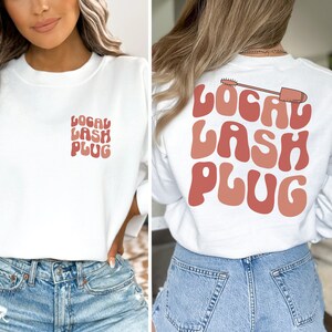 May include: White sweatshirt with the text "LOCAL LASH PLUG" in pink and brown letters on the front and back. The back also features a pink and brown mascara wand graphic.