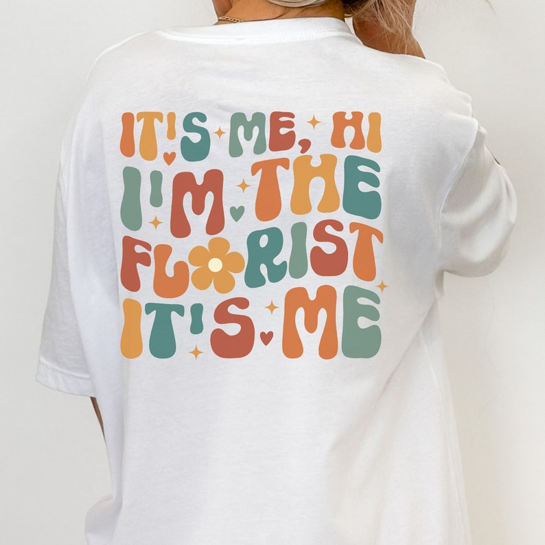 May include: White t-shirt with a retro-style graphic that says "It's me, Hi I'm the Florist It's me" in colorful, wavy letters.