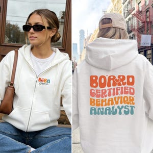 Może przedstawiać: Kremowa bluza z kapturem zapinana na zamek z napisem "BABA Jessica" z przodu i "BOARD CERTIFIED BEHAVIOR ANALYST" z tyłu w stylu retro. Bluza ma kaptur ze sznurkiem i przednie kieszenie.