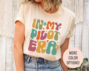 May include: A cream-colored t-shirt with the text "IN MY PILOT ERA" in colorful, retro-style lettering. The letters are outlined in black and have a 3D effect. The text is centered on the shirt.
