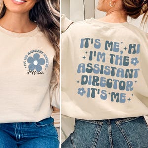 Op de afbeelding: Een beige sweatshirt met de tekst "It's me, Hi I'm the Assistant Director It's me" in blauw en wit. De tekst is in een retro-lettertype met een bloemgrafiek. Het sweatshirt wordt getoond op een model dat een blauwe jeans draagt.