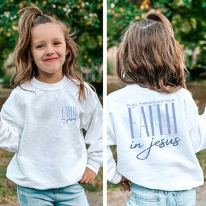 May include: White sweatshirt with the text "FAITH in Jesus" on the front and back. The front design is smaller and centered, while the back features the phrase "IN ALL THINGS PLACE YOUR FAITH in Jesus" in a larger, vertical font.