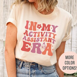 Puede incluir: Una camiseta beige claro con un gráfico de estilo retro que dice "In My Activity Assistant Era". El gráfico presenta colores rosa, naranja y morado con un diseño de mariposa.