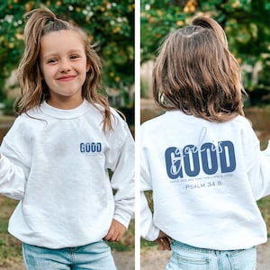 May include: White sweatshirt with the word "GOOD" printed in blue on the front and back. The back of the sweatshirt also has the phrase "GOD IS GOOD" and "TASTE AND SEE THAT THE LORD IS GOOD. PSALM 34:8" printed in blue.