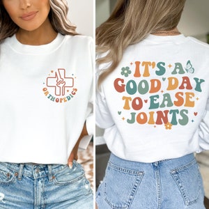 Könnte beinhalten: Weißer Pullover mit einem Grafikdruck eines Kreuzes und dem Wort "ORTHOPEDICS" auf der Vorderseite und dem Text "IT'S A GOOD DAY TO EASE JOINTS" auf der Rückseite. Die Grafik und der Text sind im Retro-Stil mit bunten Buchstaben gehalten.