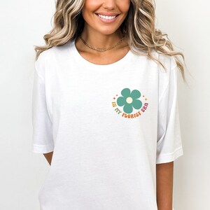 Op de afbeelding: Wit T-shirt met een groene bloemprint en de tekst "In My Florist Era" in een regenboogkleurenpalet.