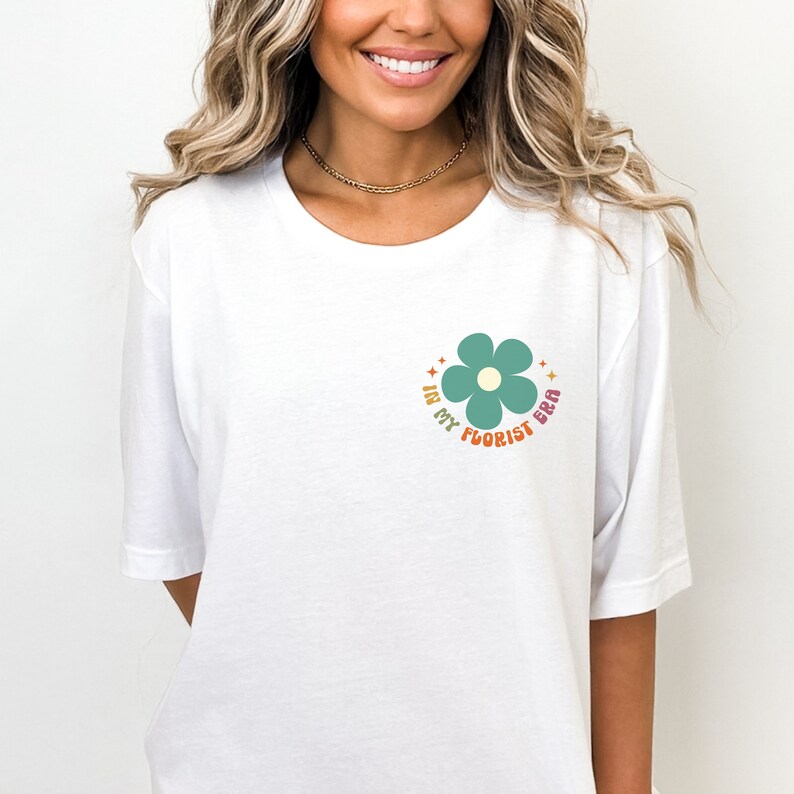 Op de afbeelding: Wit T-shirt met een groene bloemprint en de tekst "In My Florist Era" in een regenboogkleurenpalet.