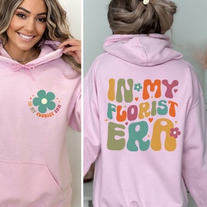 Op de afbeelding: Roze hoodie met de tekst "In My Florist Era" in kleurrijk retro-lettertype op de rug en een kleinere versie van de tekst en een bloemgrafiek op de voorkant.