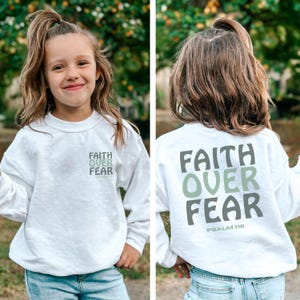 May include: White sweatshirt featuring the text "FAITH OVER FEAR" in green and grey. The sweatshirt is shown from the front and back, with the text also on the front. The text "PSALM 118" is below the text on the back.