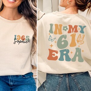 May include: Cream-colored sweatshirt with the text "1965 Sophie" on the front. The back of the sweatshirt features the words "IN MY 61 ERA" in a retro font, with butterfly and star graphics. The sweatshirt is a casual, vintage-inspired garment.