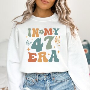 以下が含まれることがあります： 蝶と星が描かれたカラフルなレトロなフォントで「In My 47 Era」と書かれた白いスウェットシャツ。