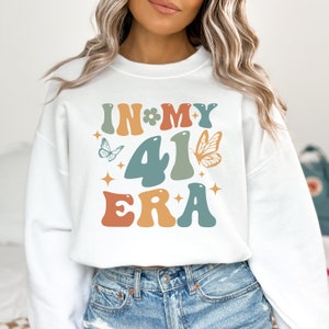 Puede incluir: Sudadera blanca con el texto "IN MY 41 ERA" en una fuente retro colorida con mariposas y estrellas.