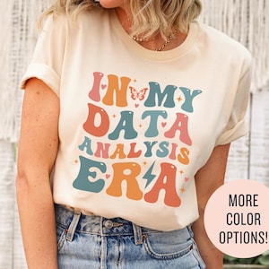 May include: A cream-coloured t-shirt with the text "In My Data Analysis Era" in a retro font with colourful accents. The text is surrounded by small hearts and a butterfly.