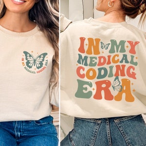 May include: A beige sweatshirt with the text "In My Medical Coding Era" in colourful retro font with a butterfly graphic. The text is on the back of the sweatshirt and the butterfly is on the front.
