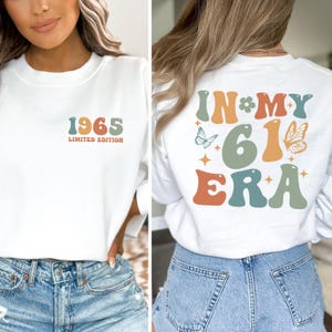 May include: White sweatshirt with the text "1965 LIMITED EDITION" on the front in retro colors. The back of the sweatshirt says "IN MY 61 ERA" with butterfly and star graphics. A casual, vintage-inspired design.