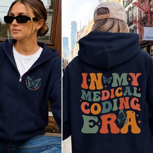 Peut inclure: Un sweat à capuche zippé bleu marine avec un petit motif de papillon sur la poitrine gauche et un grand imprimé au dos portant le texte "IN MY MEDICAL CODING ERA" en lettres rétro orange et vert sauge, agrémenté de petites étoiles et papillons.