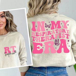 May include: A beige sweatshirt with pink lettering that reads "In My Respiratory Therapist Era" with a butterfly graphic. The sweatshirt has a small pink butterfly graphic and the text "RT Sophia" on the front.