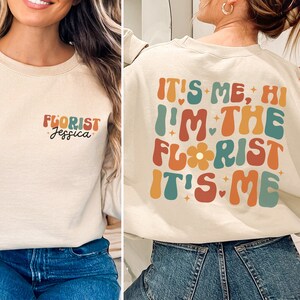 May include: A beige sweatshirt with the text "It's me, Hi I'm the Florist It's me" in colorful, retro-style lettering on the back. The front of the sweatshirt has the text "Florist Jessica" in black lettering.