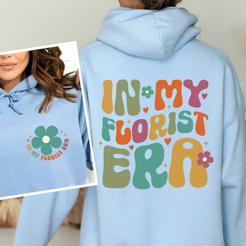 Op de afbeelding: Lichtblauwe hoodie met een grafische print van de tekst "In My Florist Era" in kleurrijke, retro-stijl letters. De grafiek bevat ook een klein bloemetjesontwerp.
