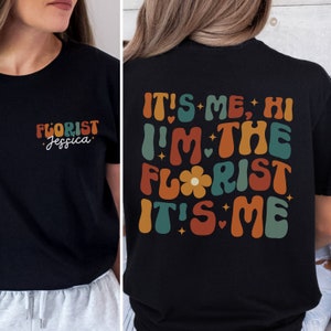 May include: Black t-shirt with a retro-style graphic that says "It's me, Hi I'm the Florist It's me". The graphic is in shades of orange, red, green, and blue. The front of the shirt has a smaller graphic that says "Florist Jessica".
