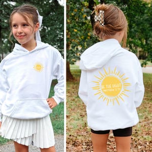 May include: White hoodie with a front pocket and hood. The front features a yellow sun graphic. The back of the hoodie displays a larger yellow sun design with the text "BE THE LIGHT MATTHEW 5:14".
