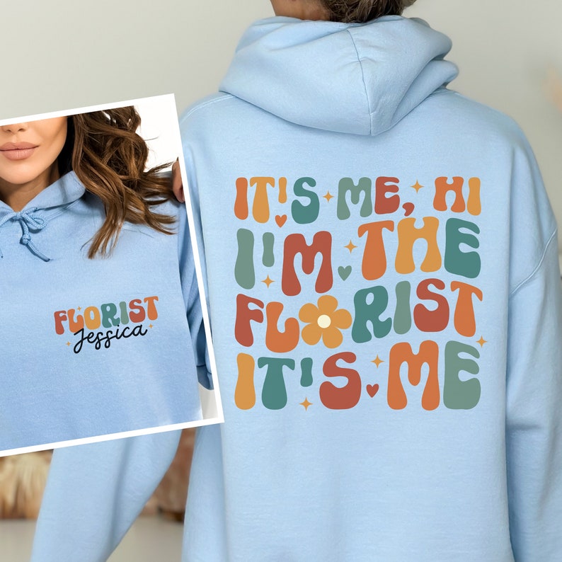 May include: Light blue hoodie with a retro-style graphic that says "It's me, hi I'm the florist it's me". The graphic is in shades of orange, red, green, and yellow. The hoodie has a drawstring hood and a kangaroo pocket.