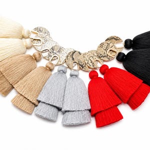May include: A collection of layered fabric tassels arranged in a semi-circle, each attached to a circular gold-toned metal charm with a snakeskin-patterned inlay. The tassels are available in cream, tan, light gray, bright red, and black colors.