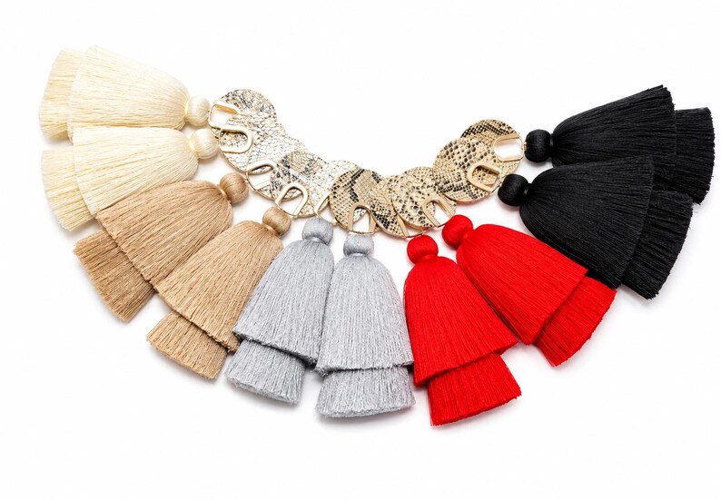 May include: A collection of layered fabric tassels arranged in a semi-circle, each attached to a circular gold-toned metal charm with a snakeskin-patterned inlay. The tassels are available in cream, tan, light grey, bright red, and black colours.