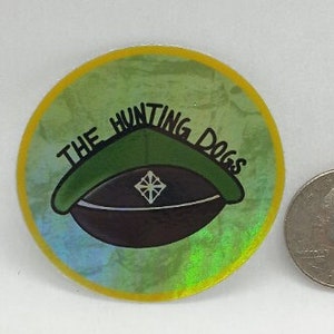 May include: A round, iridescent sticker with a yellow border. The sticker features a green military-style hat with a black brim and a white diamond shape on the front. The text "THE HUNTING DOGS" is written in black around the hat.
