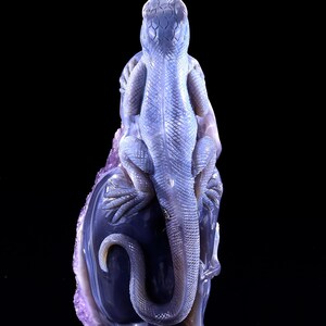 May include: A purple and blue agate carving of a lizard perched on a rock. The lizard is detailed with scales and has a long tail.