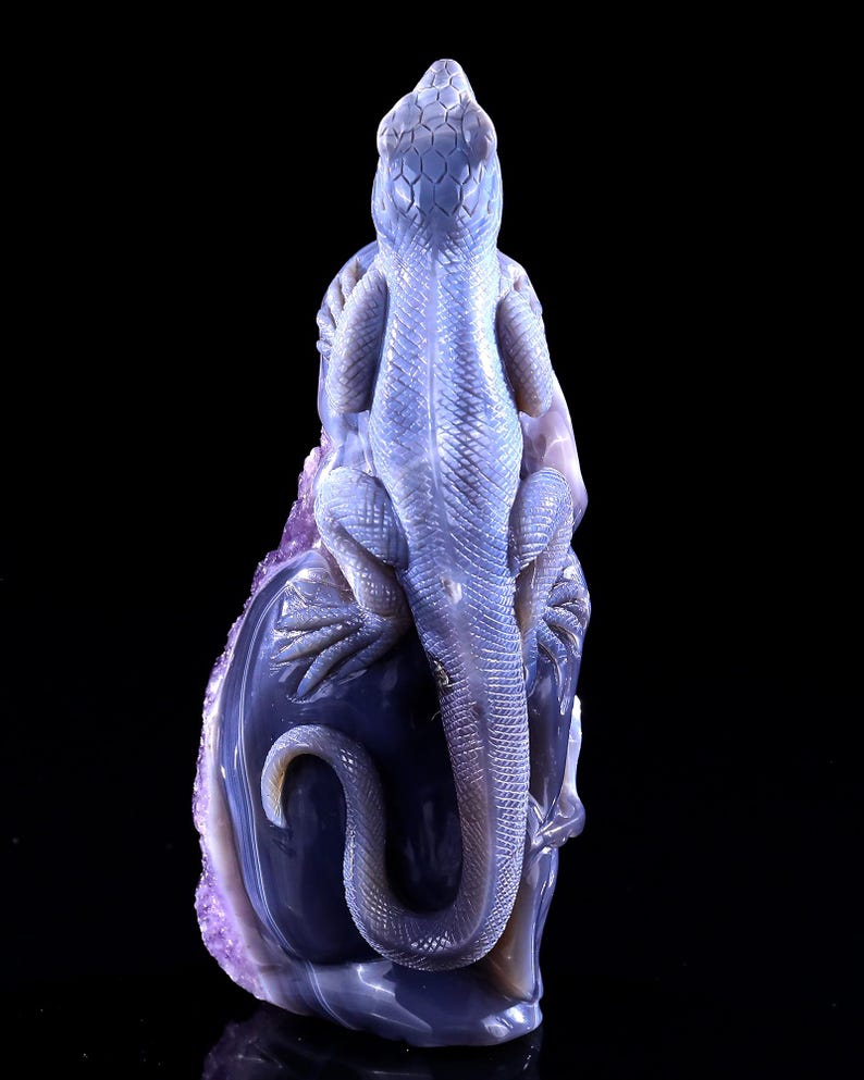 May include: A purple and blue agate carving of a lizard perched on a rock. The lizard is detailed with scales and has a long tail.