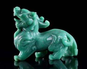 Puede incluir: Una figura de piedra verde tallada que representa una criatura mítica con cabeza de león, boca abierta y melena fluida. La escultura presenta intrincados patrones en espiral a lo largo de su cuerpo, una cola enroscada y una superficie lisa.
