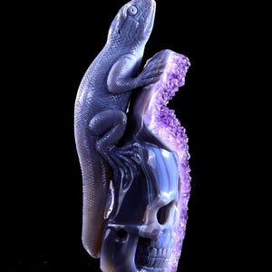 May include: A purple and grey agate skull sculpture with a carved lizard perched on top. The skull is detailed with a carved face and the lizard has a detailed body and tail.