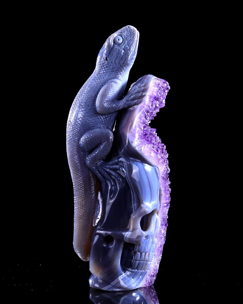 May include: A purple and grey agate skull sculpture with a carved lizard perched on top. The skull is detailed with a carved face and the lizard has a detailed body and tail.