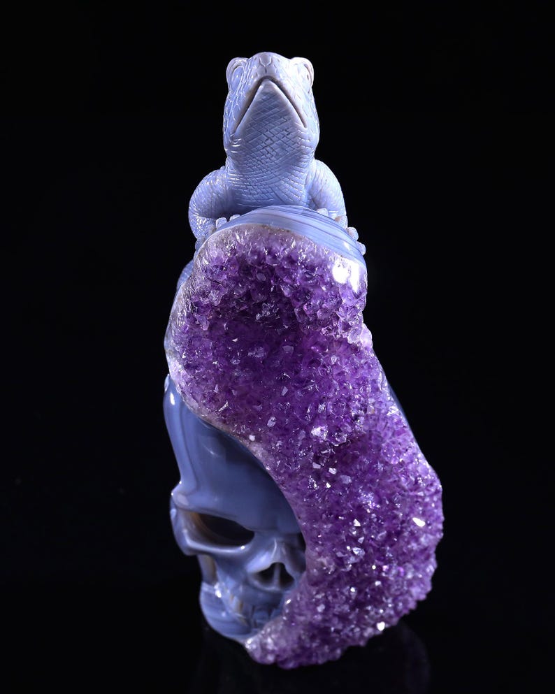 May include: A blue agate skull sculpture with a carved lizard on top. The skull is partially covered in amethyst crystals.