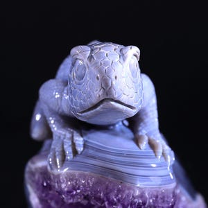 May include: A detailed carving of a lizard, crafted from light blue agate, perched atop a purple amethyst geode. The lizard's eyes are carved with a realistic detail, and its textured skin is visible.