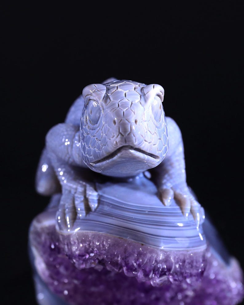 May include: A detailed carving of a lizard, crafted from light blue agate, perched atop a purple amethyst geode. The lizard's eyes are carved with a realistic detail, and its textured skin is visible.