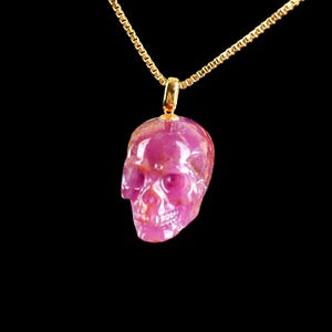 Op de afbeelding: Een roze schedel hanger hangt aan een gouden ketting tegen een zwarte achtergrond. De schedel is gesneden uit een doorschijnende, roze edelsteen, met zichtbare details van de oogkassen, neus en tanden. De gouden ketting is delicaat en fijn geschakeld.