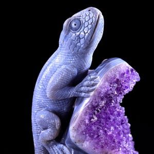 May include: A blue agate lizard sculpture perched on a purple amethyst geode. The lizard is carved in detail and has a realistic appearance. The amethyst geode is a natural crystal formation with a beautiful purple colour.
