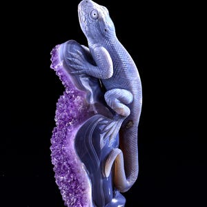 May include: A purple amethyst geode with a carved agate lizard perched on top. The lizard is facing upwards with its mouth slightly open. The geode is a natural formation with a rough, textured surface.