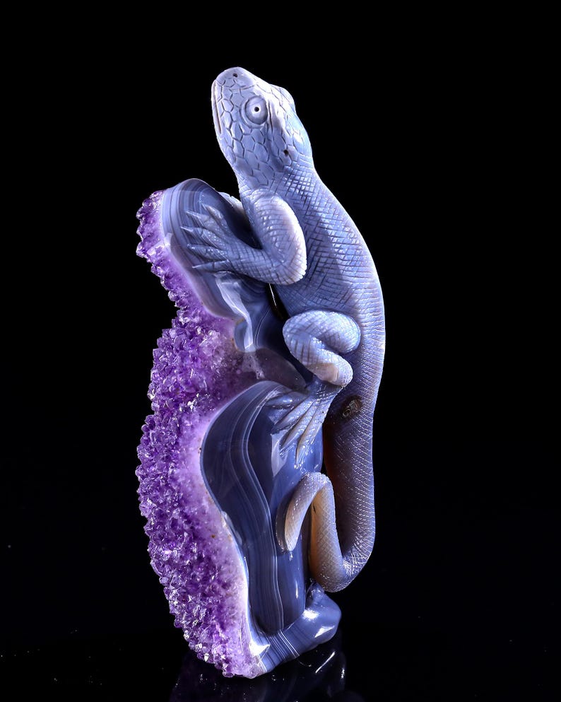 May include: A purple amethyst geode with a carved agate lizard perched on top. The lizard is facing upwards with its mouth slightly open. The geode is a natural formation with a rough, textured surface.
