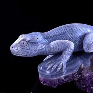 May include: A blue agate lizard sculpture perched on a purple amethyst base. The lizard is carved in detail, with scales and claws visible. The amethyst base is rough and textured.