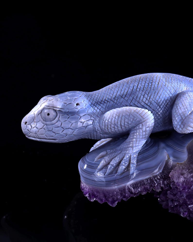 May include: A blue agate lizard sculpture perched on a purple amethyst base. The lizard is carved in detail, with scales and claws visible. The amethyst base is rough and textured.