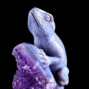 May include: A blue agate lizard sculpture perched on a purple amethyst geode. The lizard is carved in detail and has a textured surface. The amethyst geode is a natural crystal formation with a rough, jagged edge.