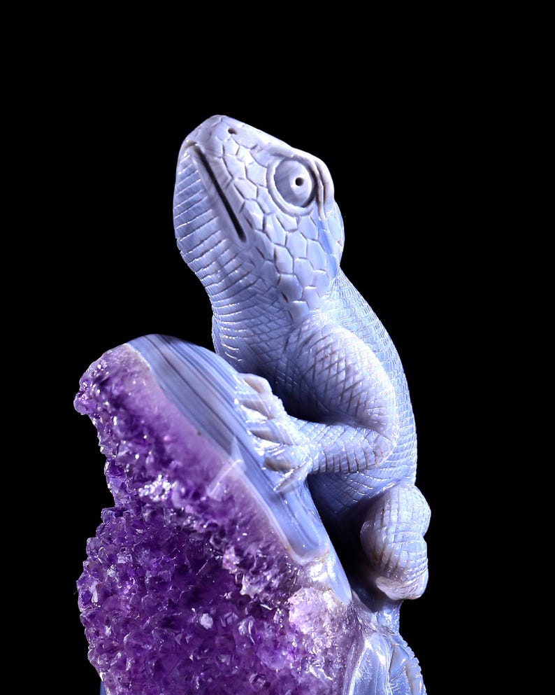 May include: A blue agate lizard sculpture perched on a purple amethyst geode. The lizard is carved in detail and has a textured surface. The amethyst geode is a natural crystal formation with a rough, jagged edge.